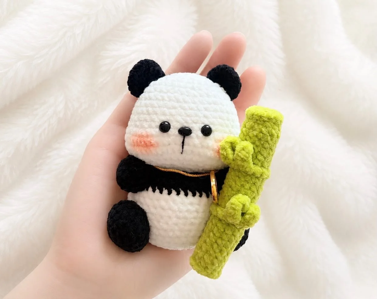 Seated black and white crochet panda holding a large green bamboo stalk, resting in a hand over a white blanket