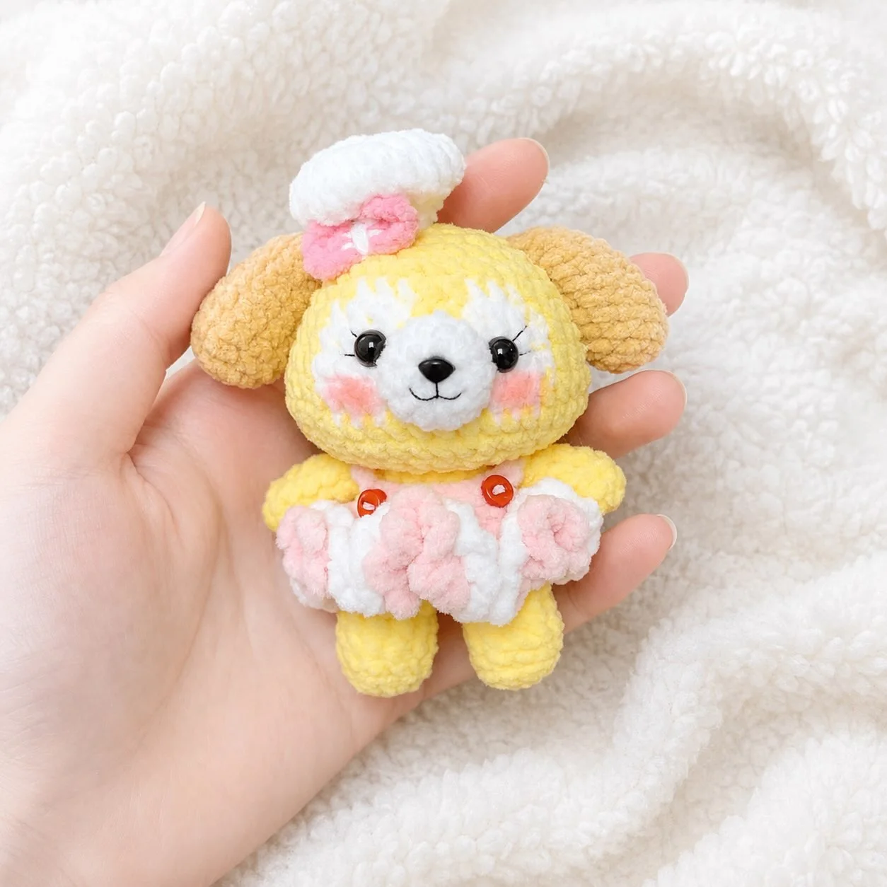 Yellow crochet dog wearing a white hat with a pink bow and a pink and white outfit, held over a white blanket