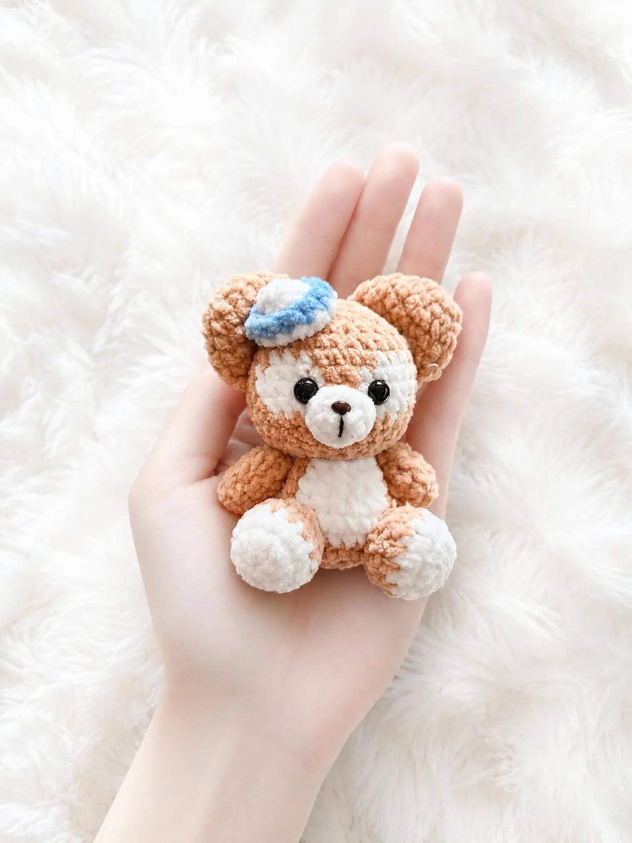 Brown crochet bear with a cream muzzle and tummy and a small blue hat, held over a white blanket