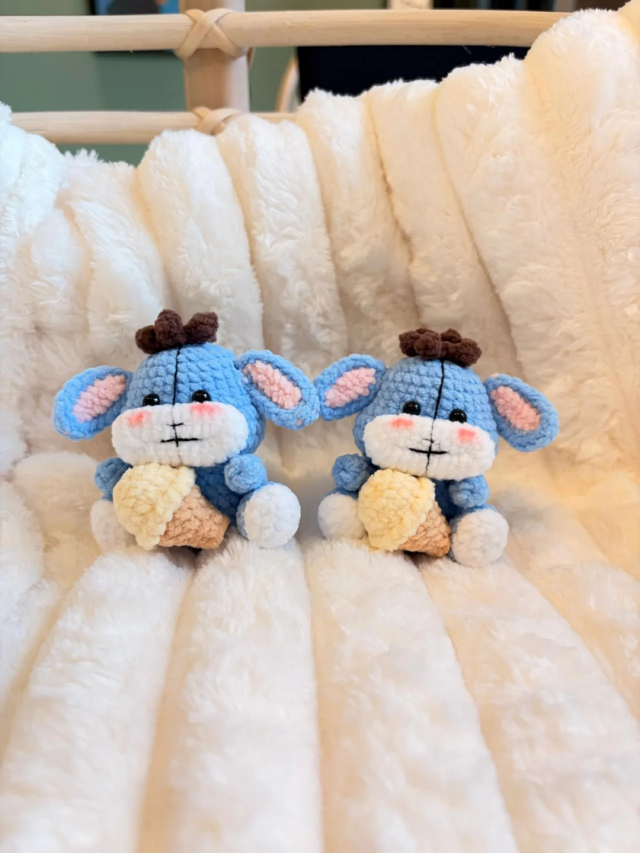 Two blue crochet animal companions with large pink-lined ears, brown tufts and cream tummies, sitting on a white blanket