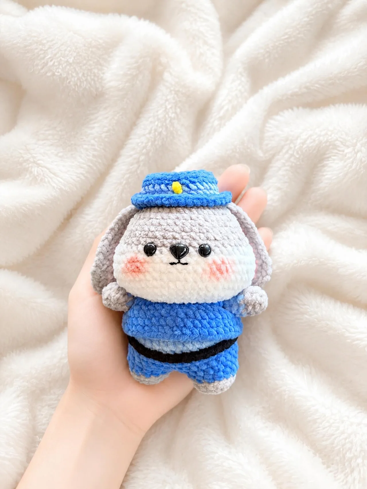 Grey floppy-eared crochet dog in a blue uniform and matching cap, resting in a hand over a white blanket