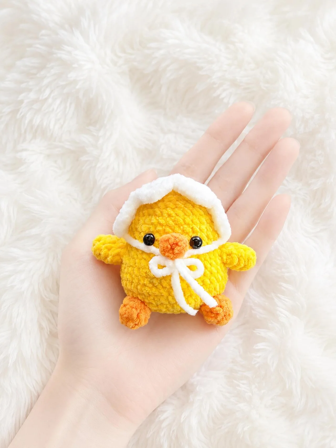 Yellow crochet duckling wearing a white bonnet tied beneath its beak, resting in a hand over a white blanket