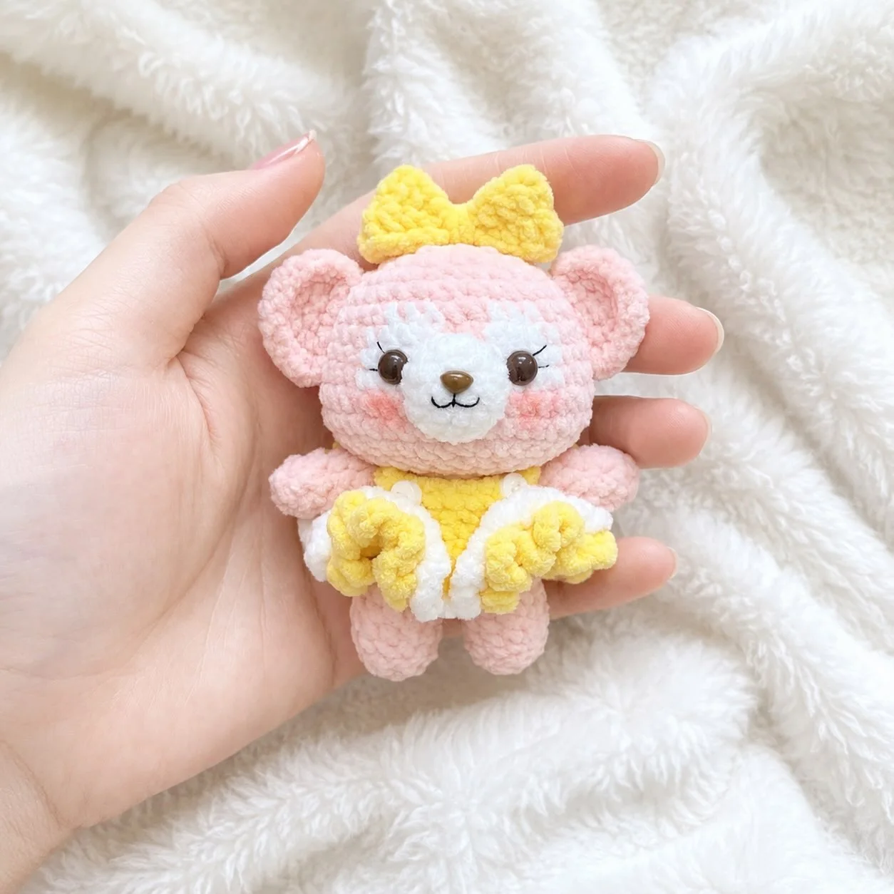 Pink crochet bear with a yellow bow and a yellow and white outfit, held over a white blanket