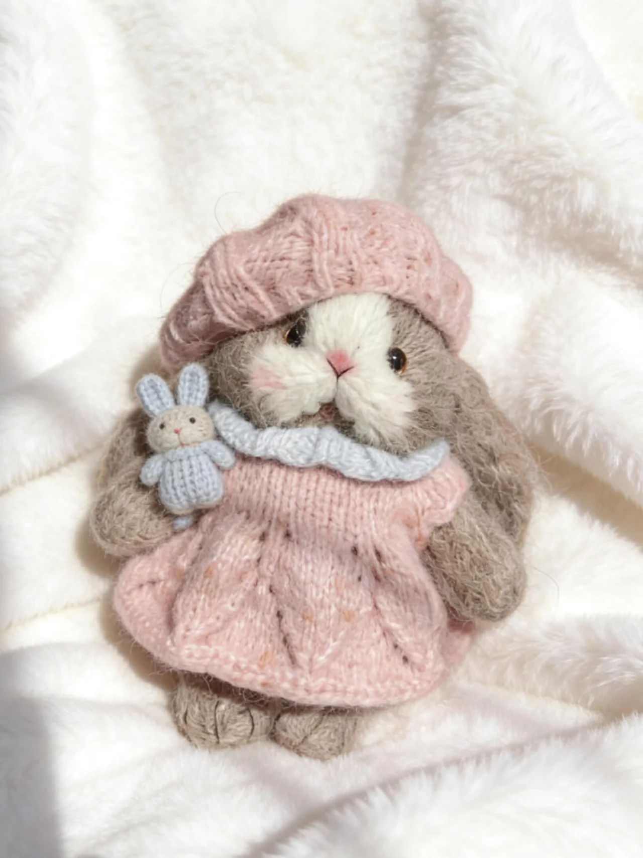 Soft knitted bunny in a pink dress and matching hat, holding a tiny blue bunny on a white blanket