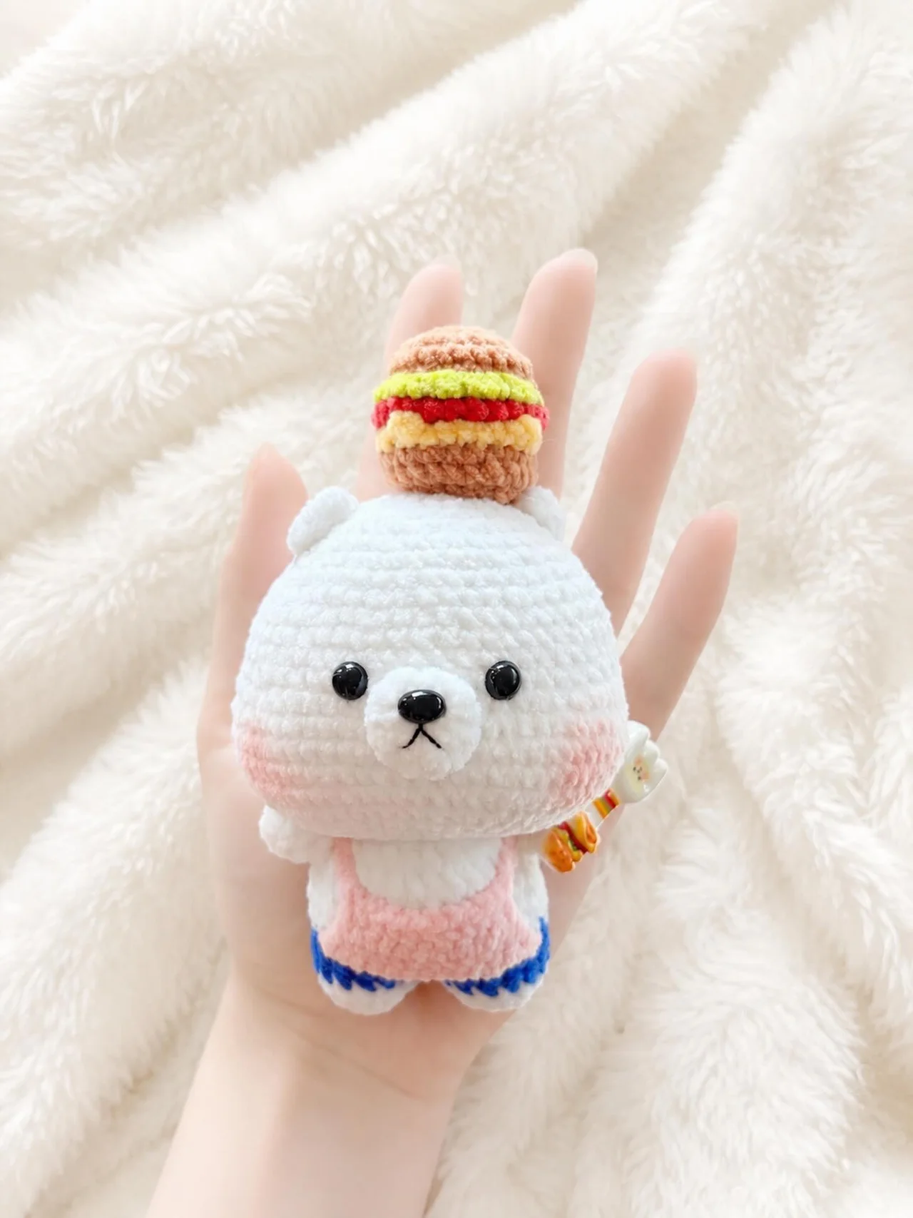 White crochet bear with a tiny burger hat, pink dungarees and blue trim, held over a white blanket