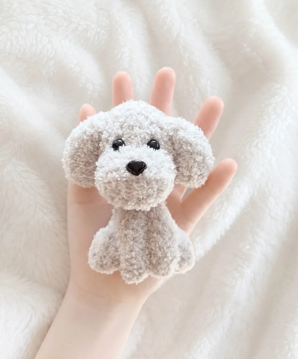 Soft grey crochet dog with floppy ears, a cream muzzle and black nose, held over a white blanket