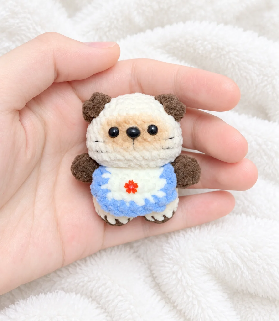 Tiny cream crochet Siamese cat with brown ears and paws, a tan face and a blue-edged bib with a red flower, resting in a hand