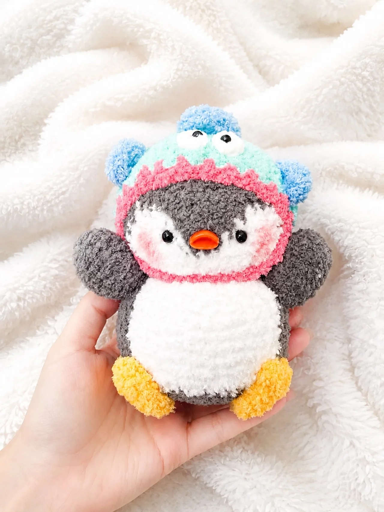 Grey and white crochet penguin in a blue dinosaur hood with a pink opening, held over a white blanket