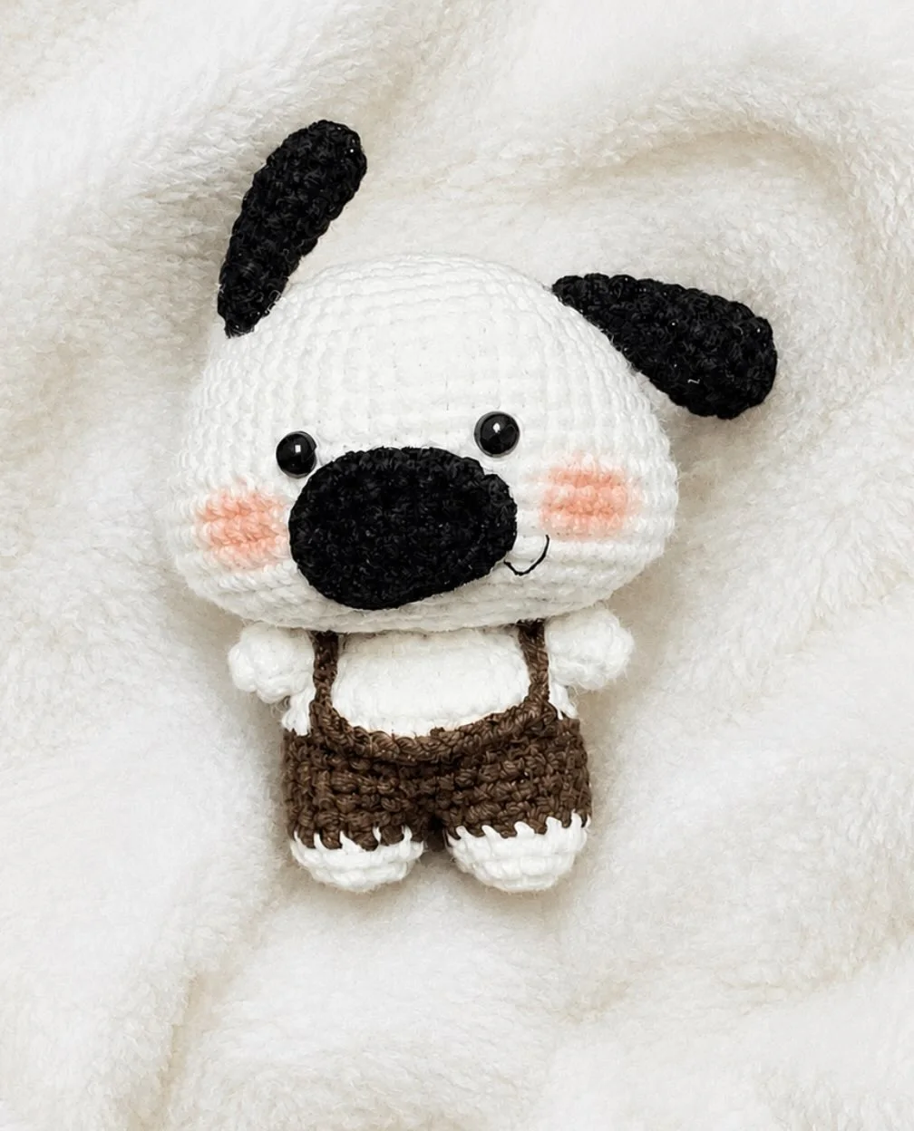 White crochet dog with black ears and a large black nose, wearing brown dungarees on a white blanket