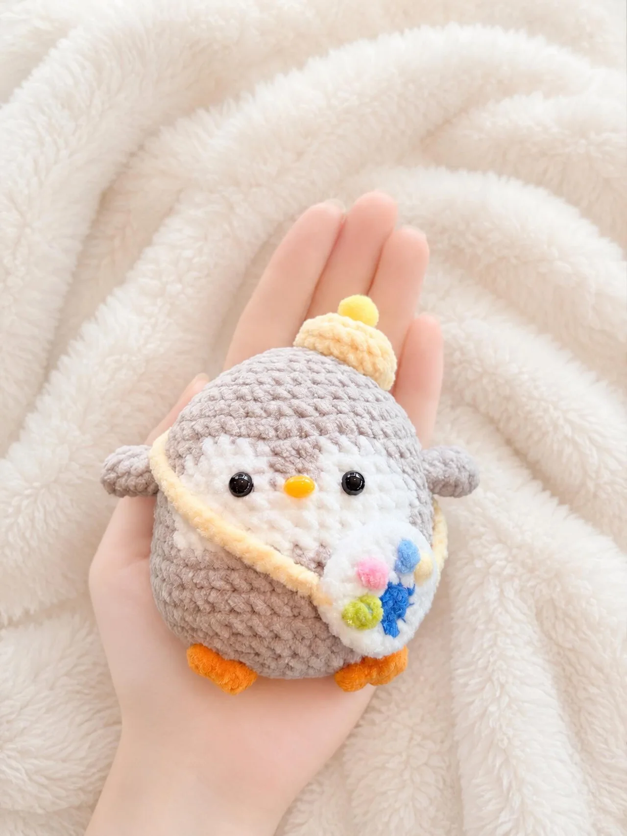 Grey crochet penguin with orange feet, a little yellow hat and a cream shoulder bag decorated with flowers, held over a white blanket