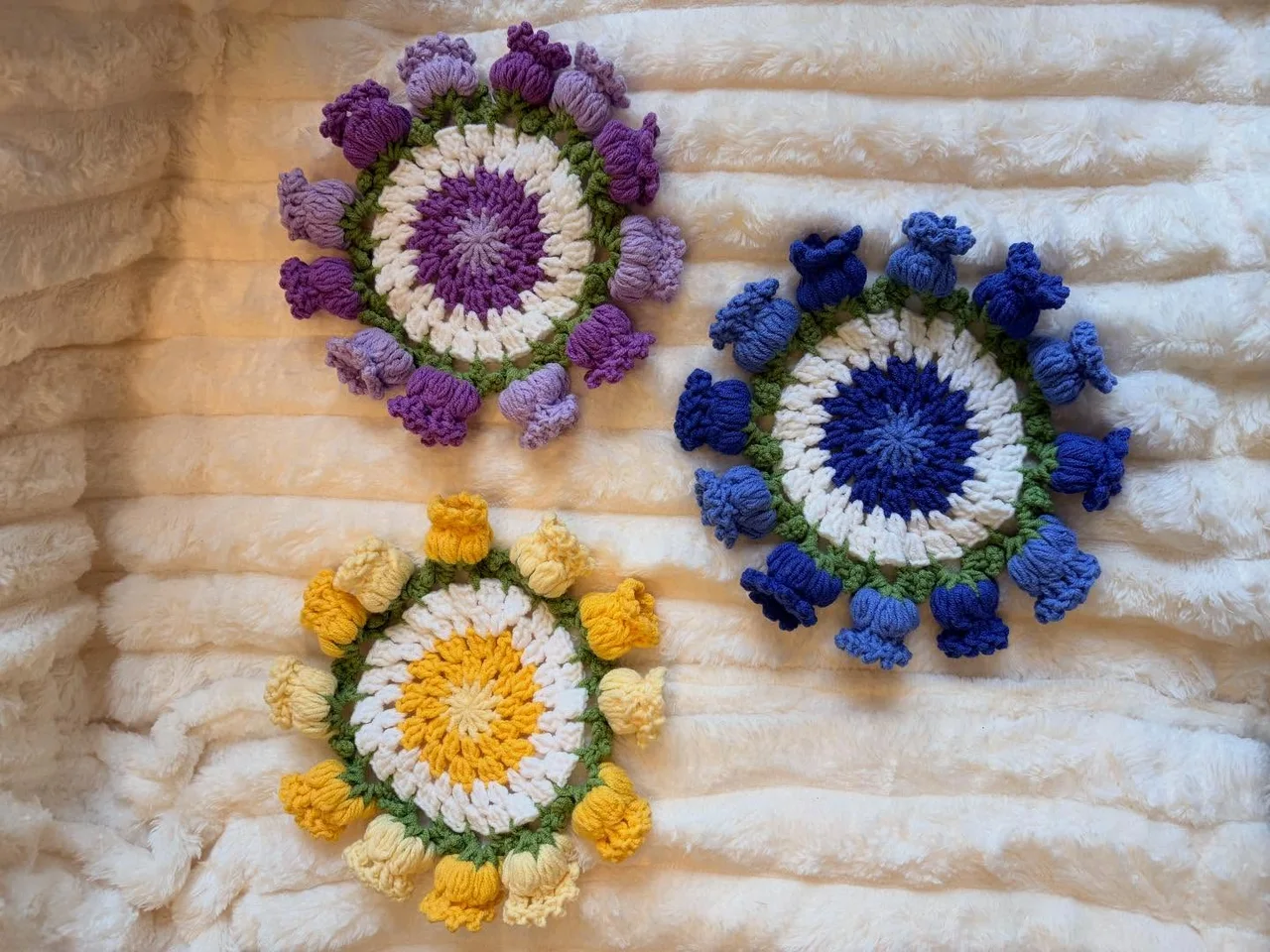 Round crochet coasters edged with raised purple, blue and yellow flowers, laid flat on a white blanket