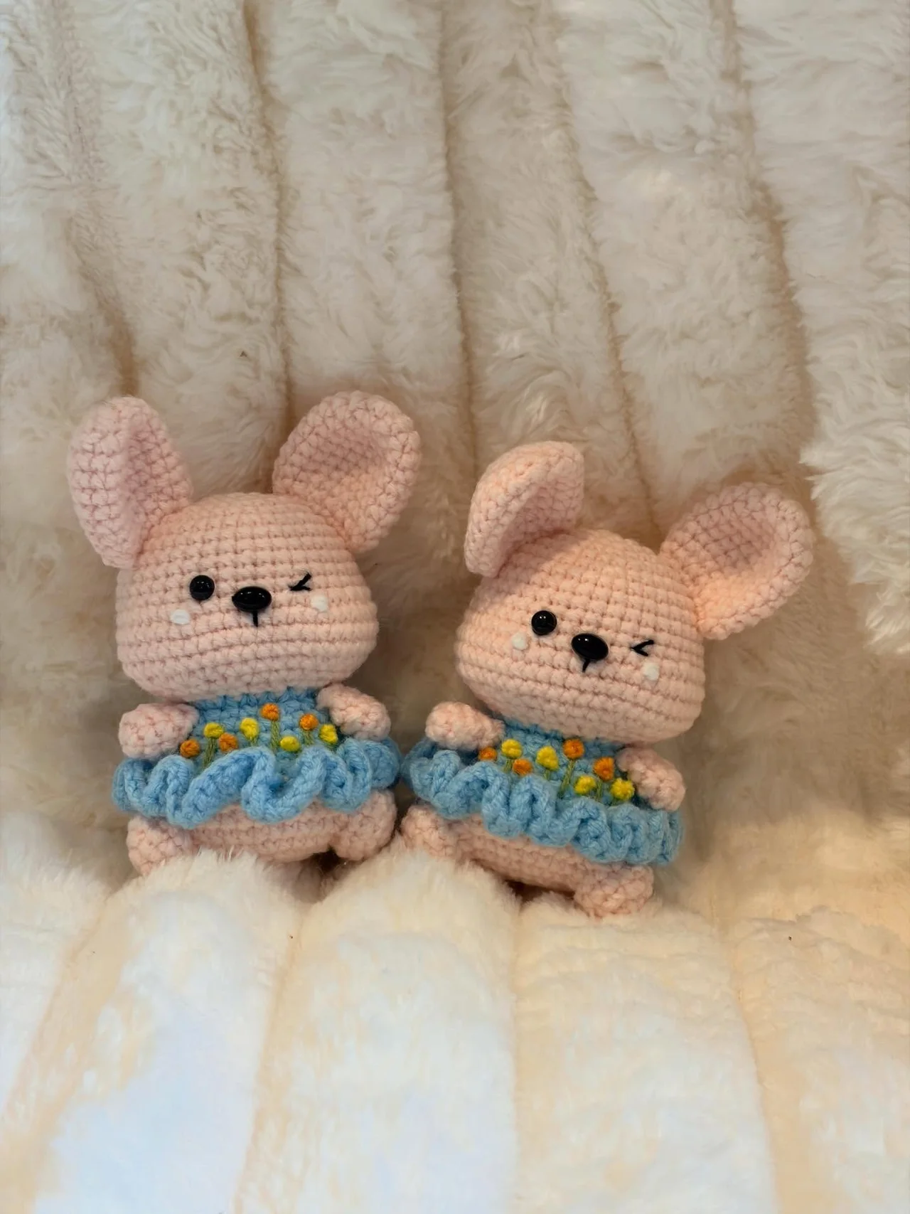 Two pink crochet bunnies wearing blue skirts edged with tiny multicoloured flowers, sitting on a white blanket