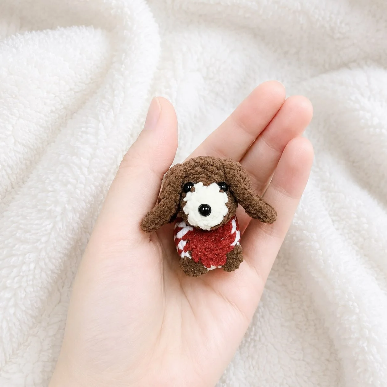 Brown floppy-eared crochet dog with a white muzzle and a red and white jumper, resting in a hand over a white blanket