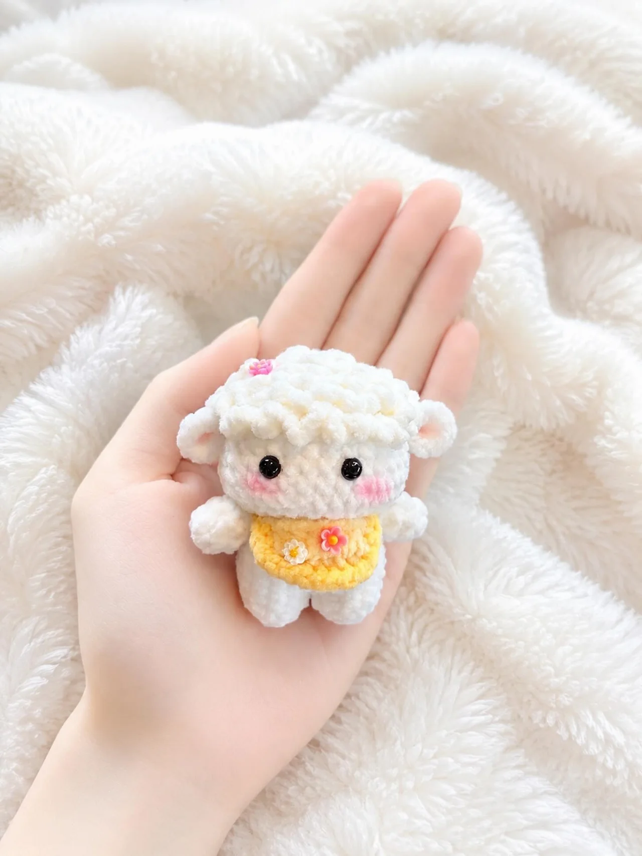 Small white crochet lamb with curly fleece and a yellow floral bib, resting in a hand over a white blanket