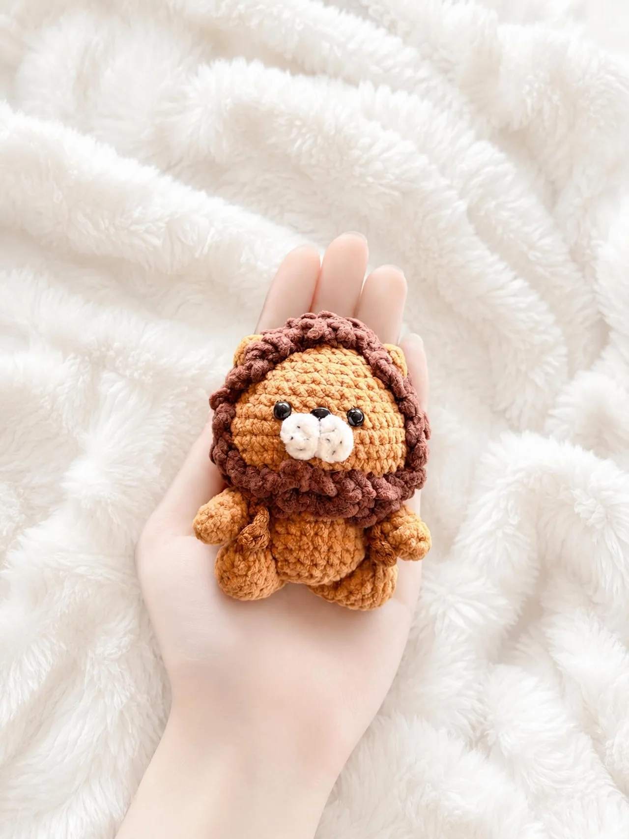 Small golden crochet lion with a soft brown mane and rounded paws, resting in a hand over a white blanket