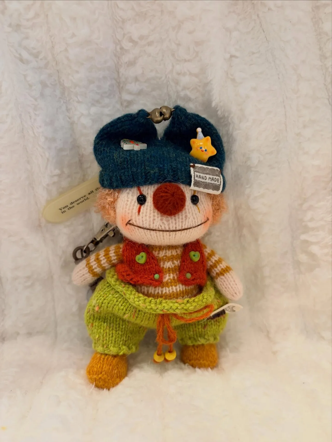 Knitted clown in a blue hat and bright green trousers, with colourful buttons and a round red nose, sitting on a white blanket