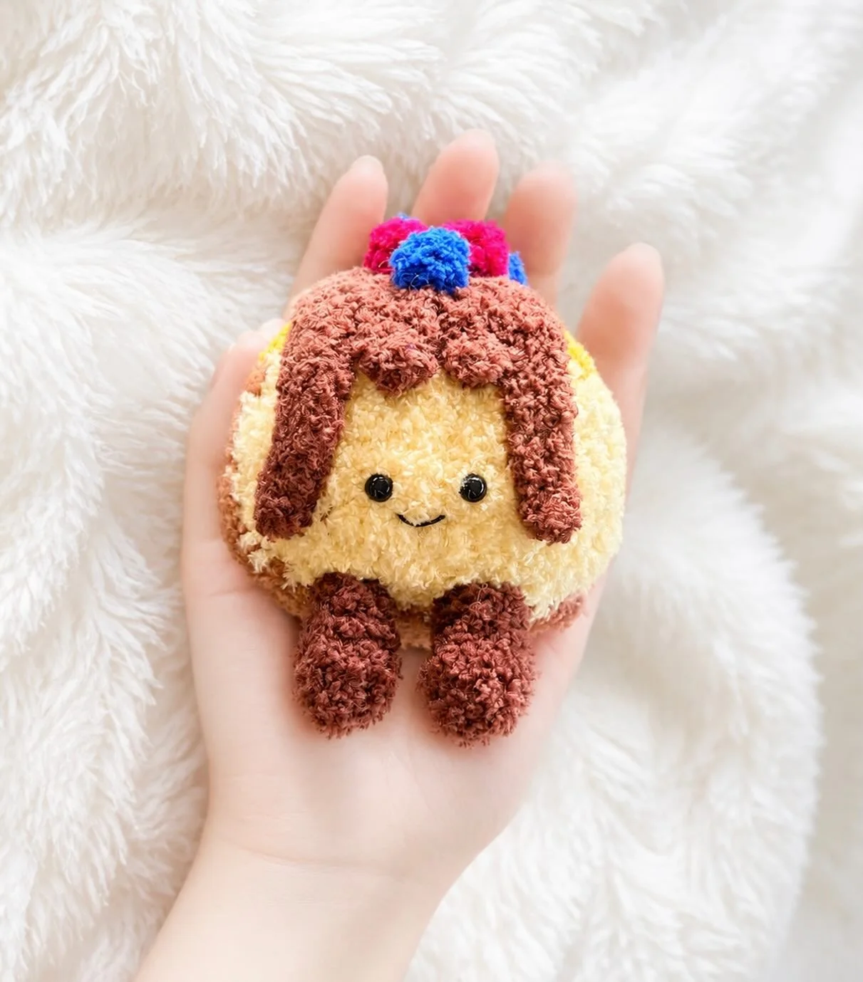Smiling golden crochet pudding with chocolate-coloured topping and dangling legs, held over a white blanket