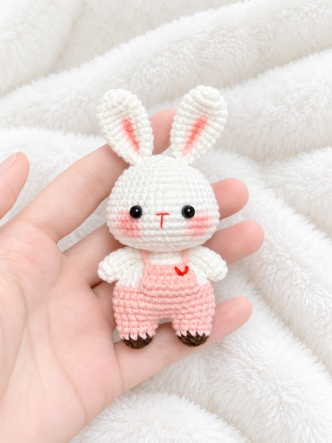 White crochet bunny with upright pink-lined ears, pink dungarees embroidered with a red heart and brown feet, resting in a hand