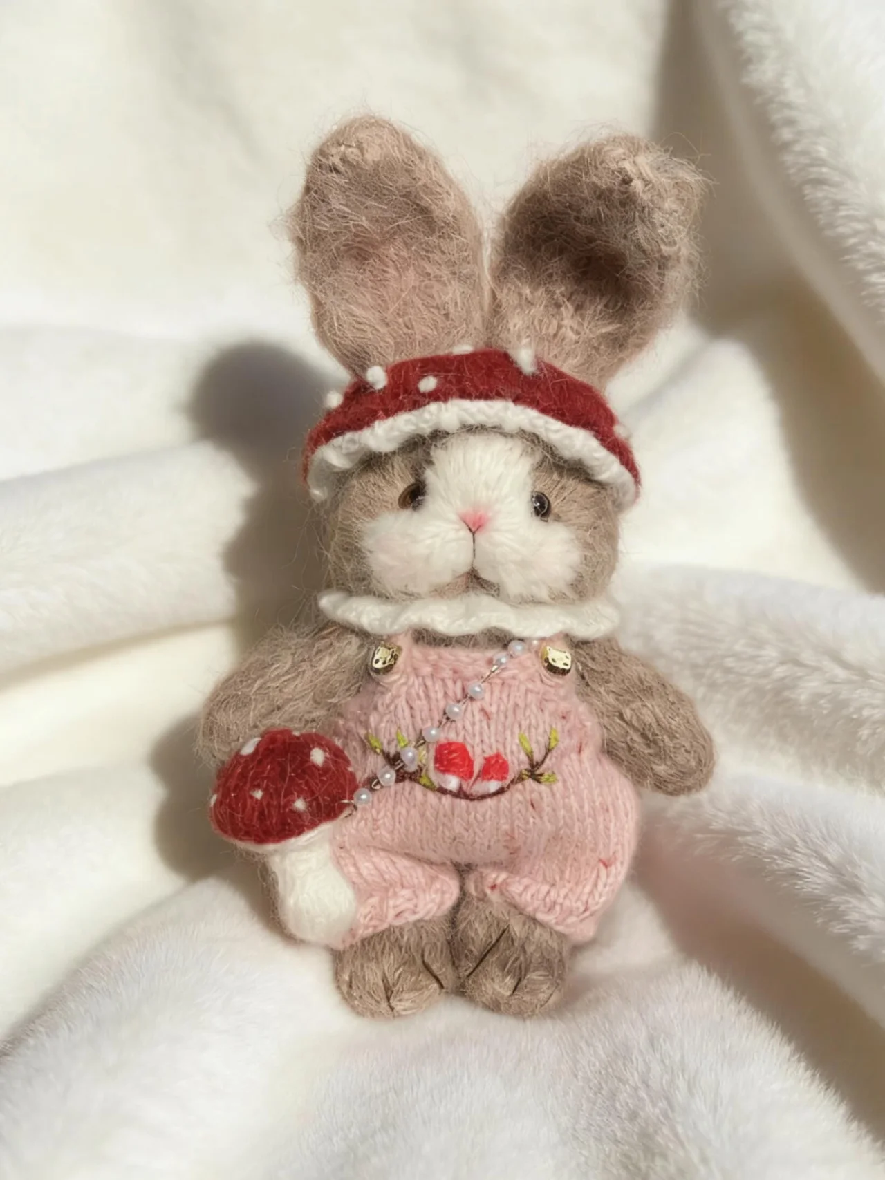 Soft knitted bunny in pink dungarees and a red mushroom headband, holding a small red mushroom on a white blanket