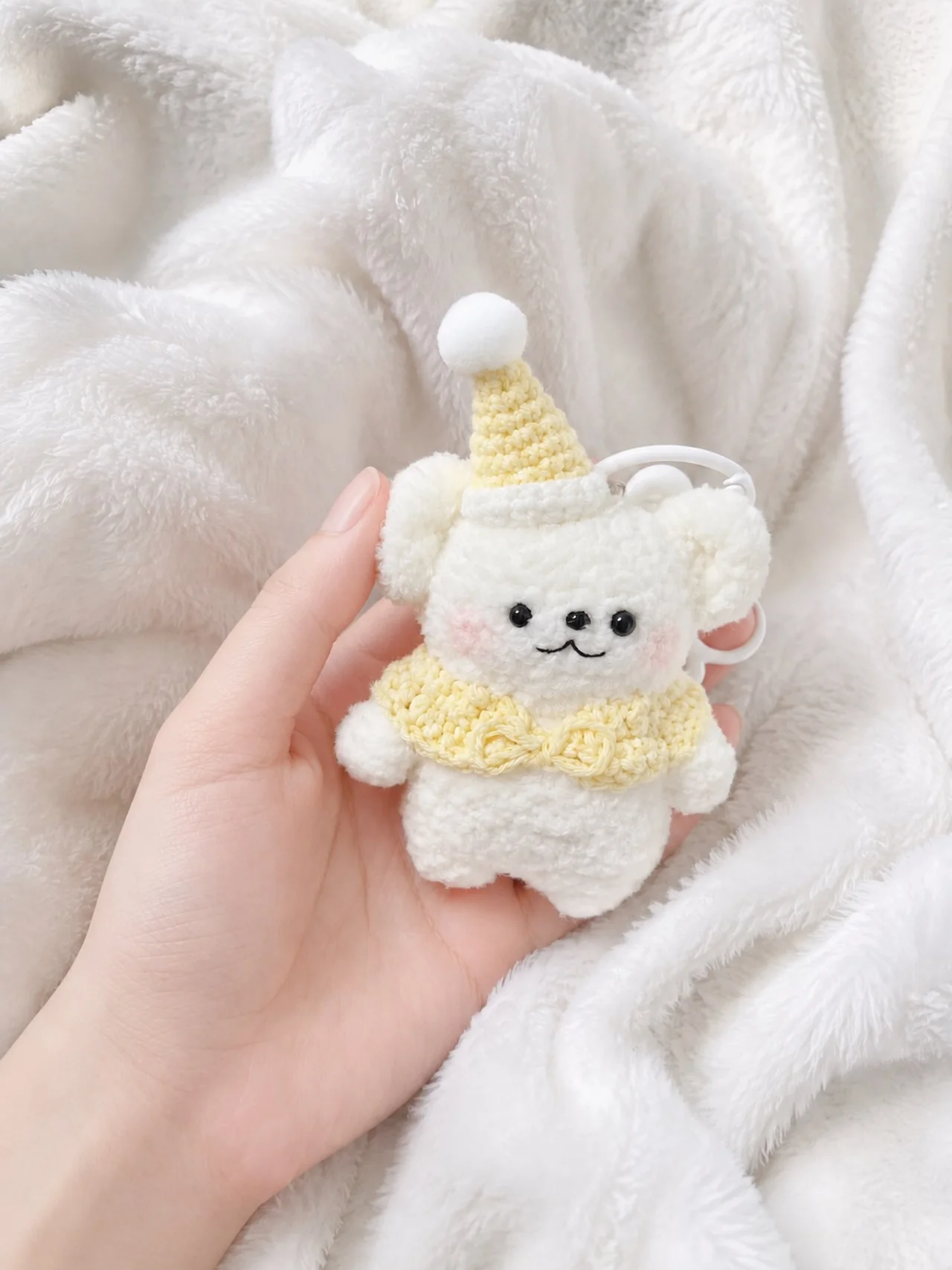 Cream crochet bear with a pale yellow pointed party hat and matching ruffled collar, held over a white blanket