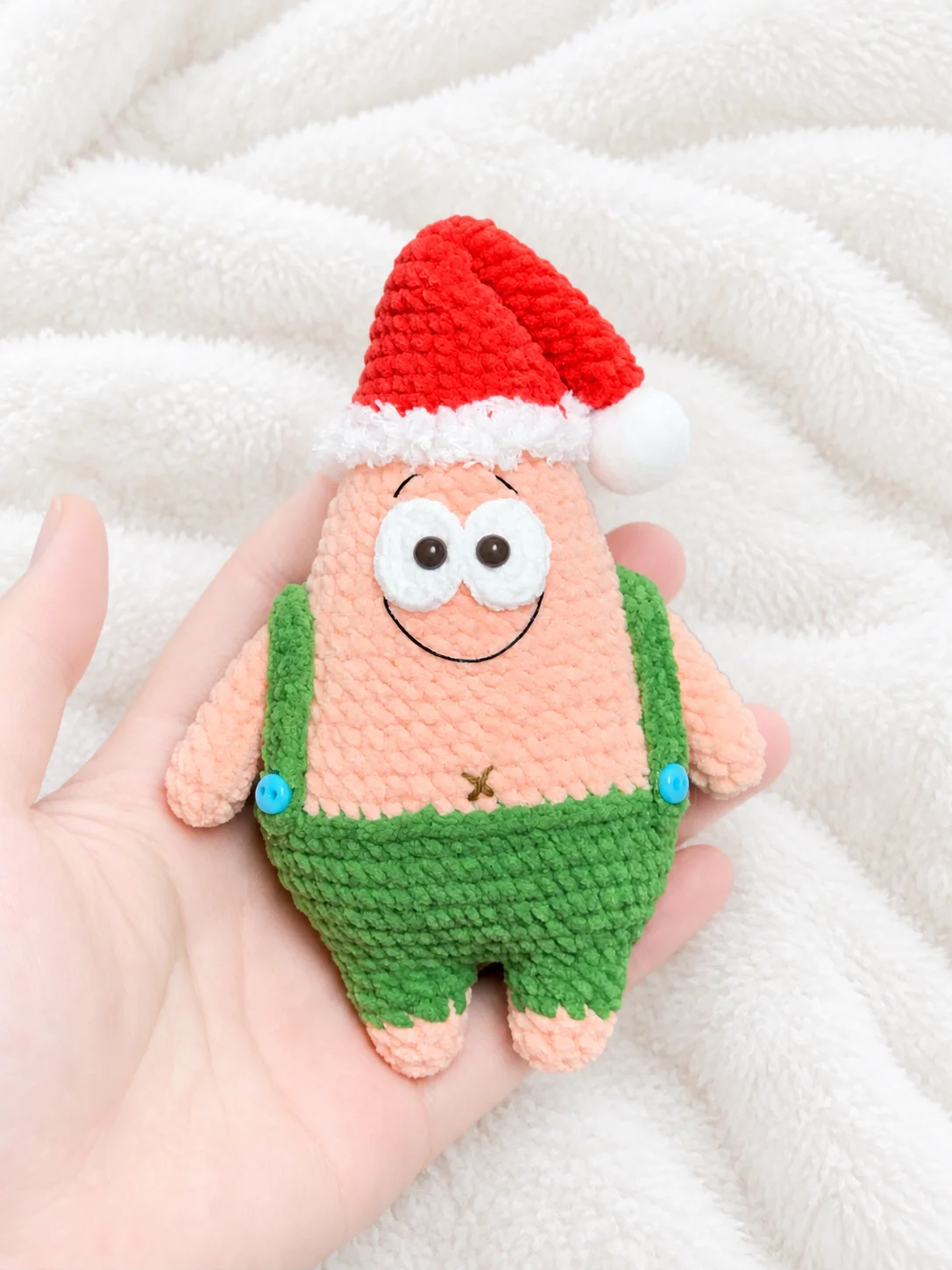 Pink crochet Patrick Star wearing a red Santa hat with white trim and a pom-pom, green shorts and braces with blue buttons, resting in a hand