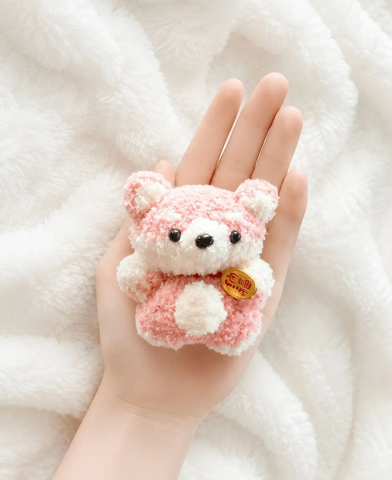 Fluffy pink and cream crochet fox with pointed ears and a small gold charm, resting in a hand over a white blanket