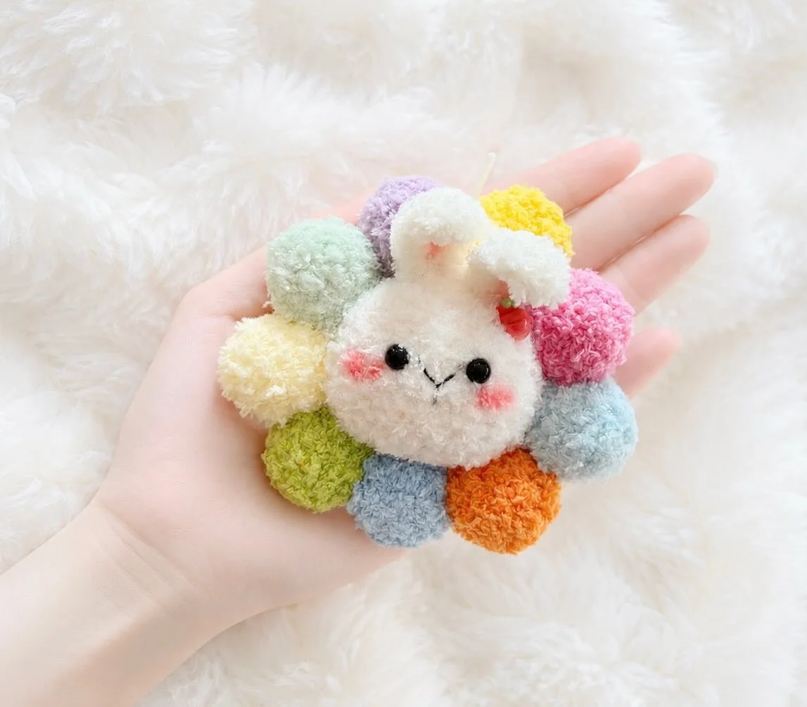 Cream bunny face and ears surrounded by fluffy rainbow petals, held over a white blanket