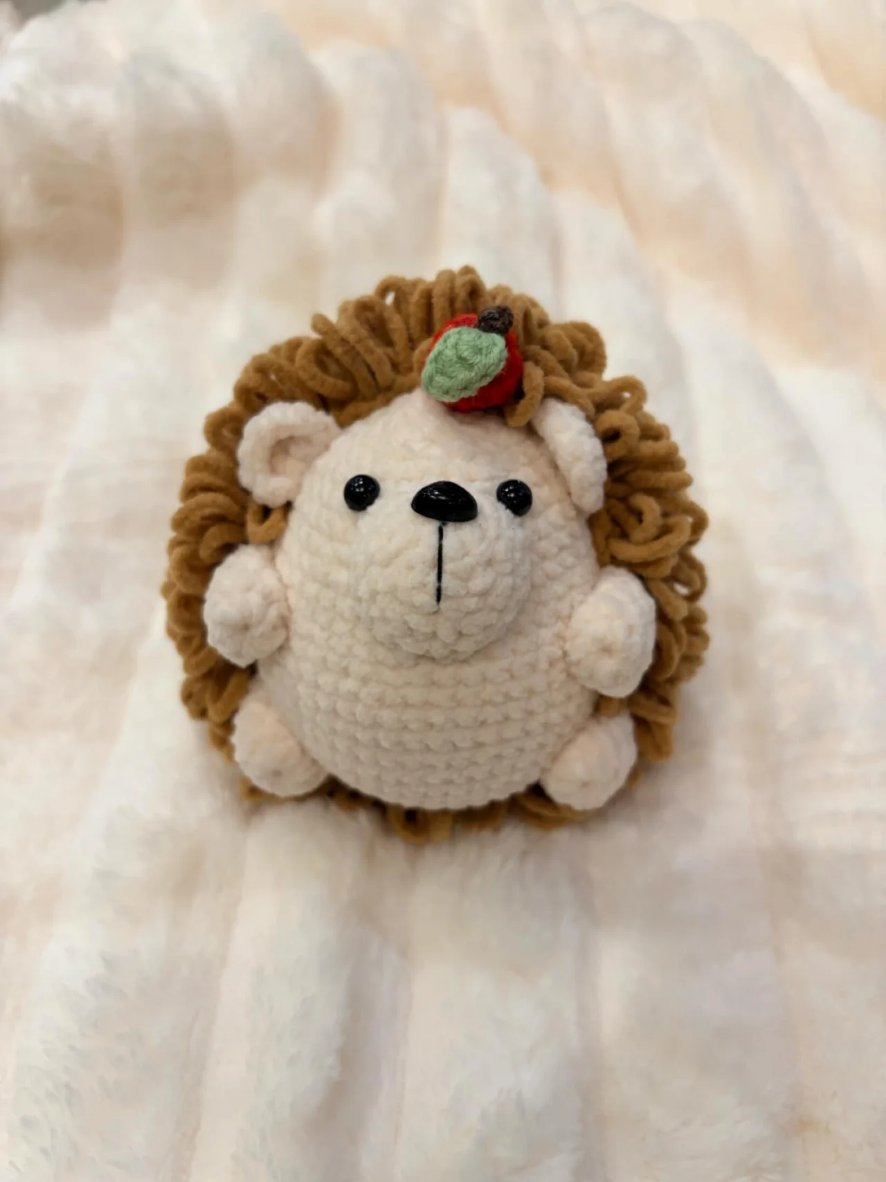 Round cream crochet hedgehog with brown looped spines and a little green leaf detail, sitting on a white blanket