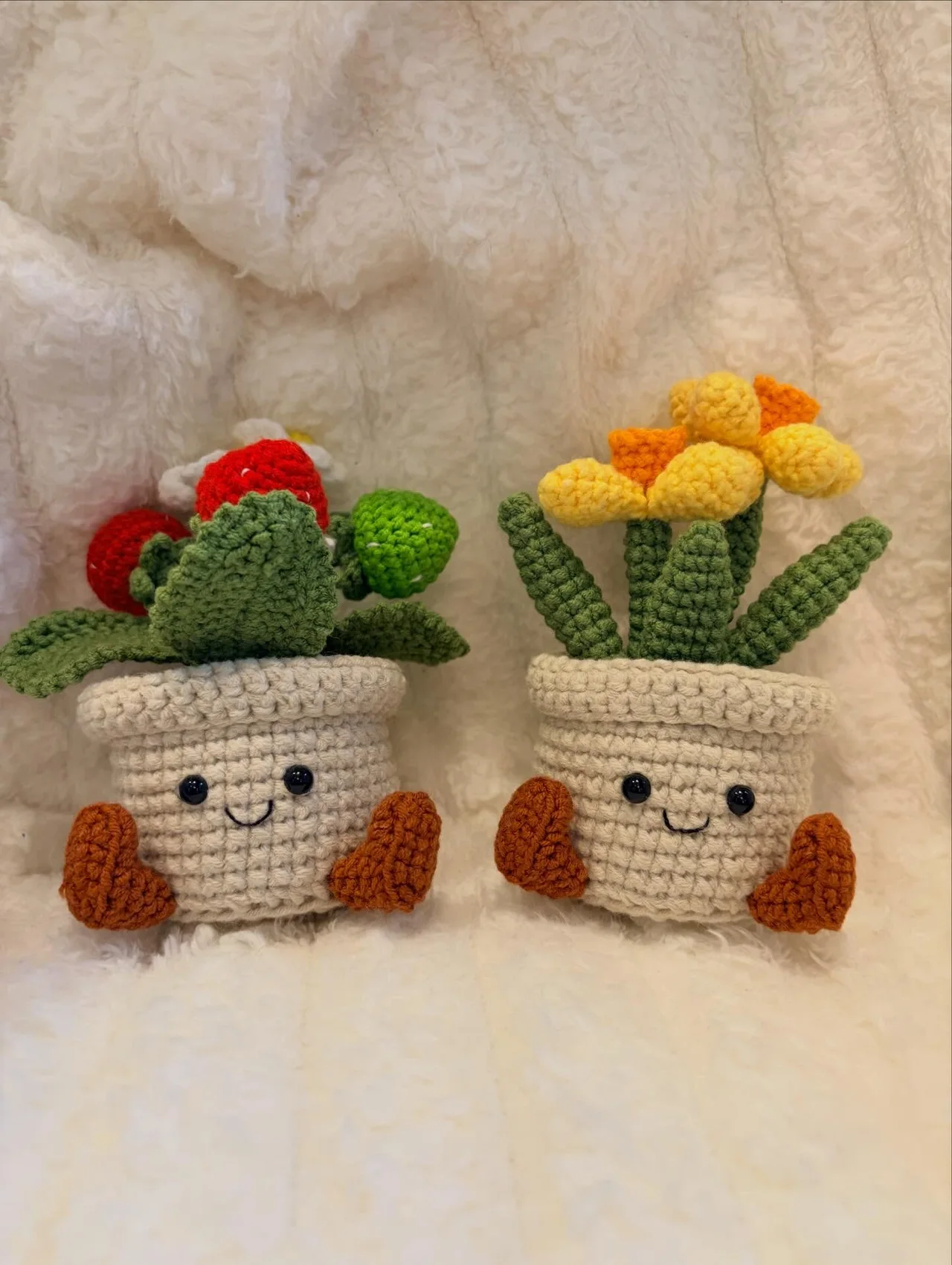 Two smiling cream crochet plant pots with little brown feet, one holding a cactus and one holding yellow flowers, on a white blanket