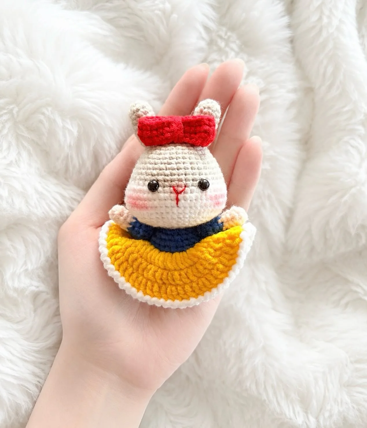 White crochet bunny dressed as Snow White, with a red bow, blue bodice and bright yellow skirt, resting in a hand