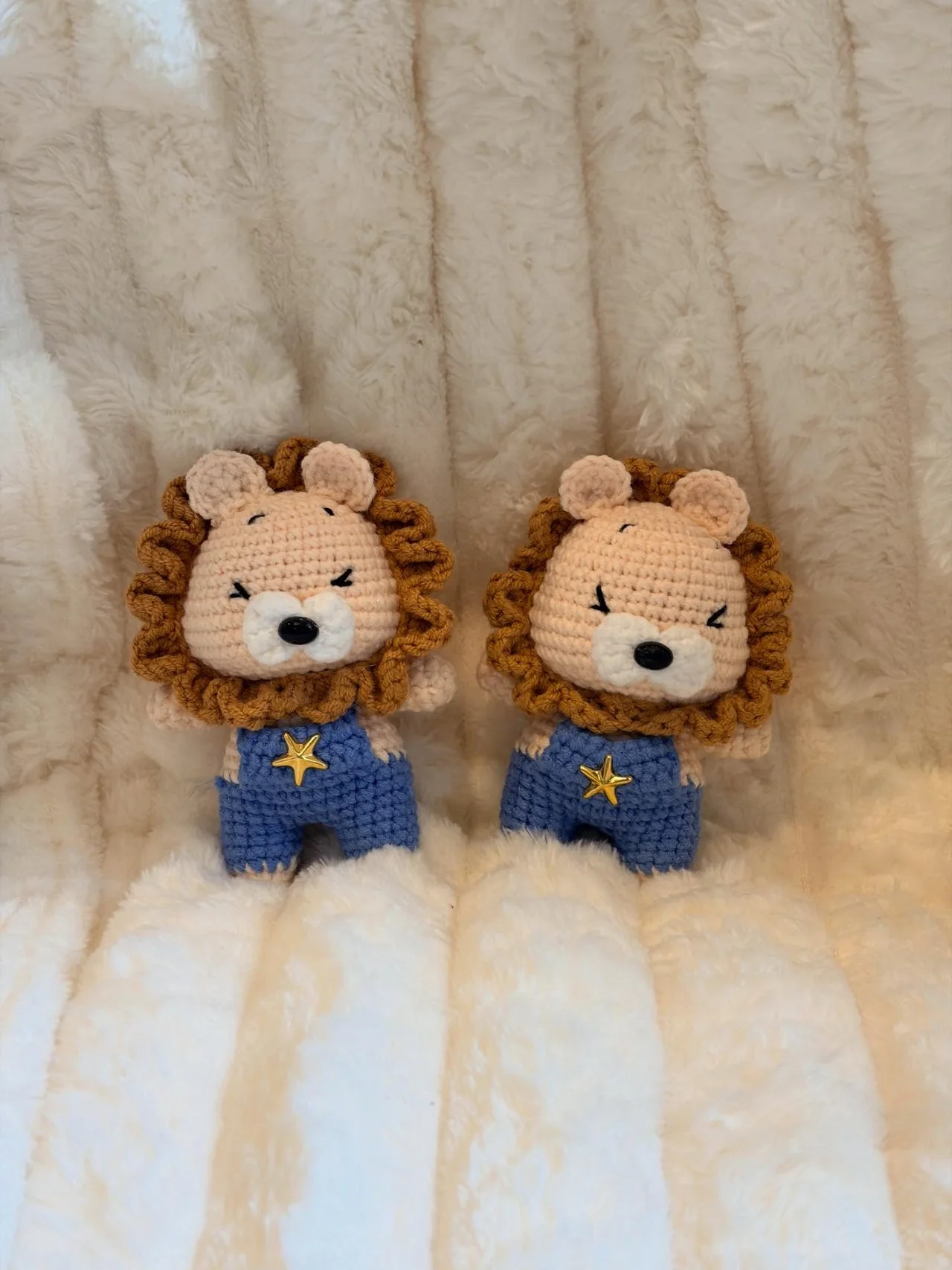 Two crochet lions with brown manes in blue dungarees decorated with yellow stars, sitting on a white blanket