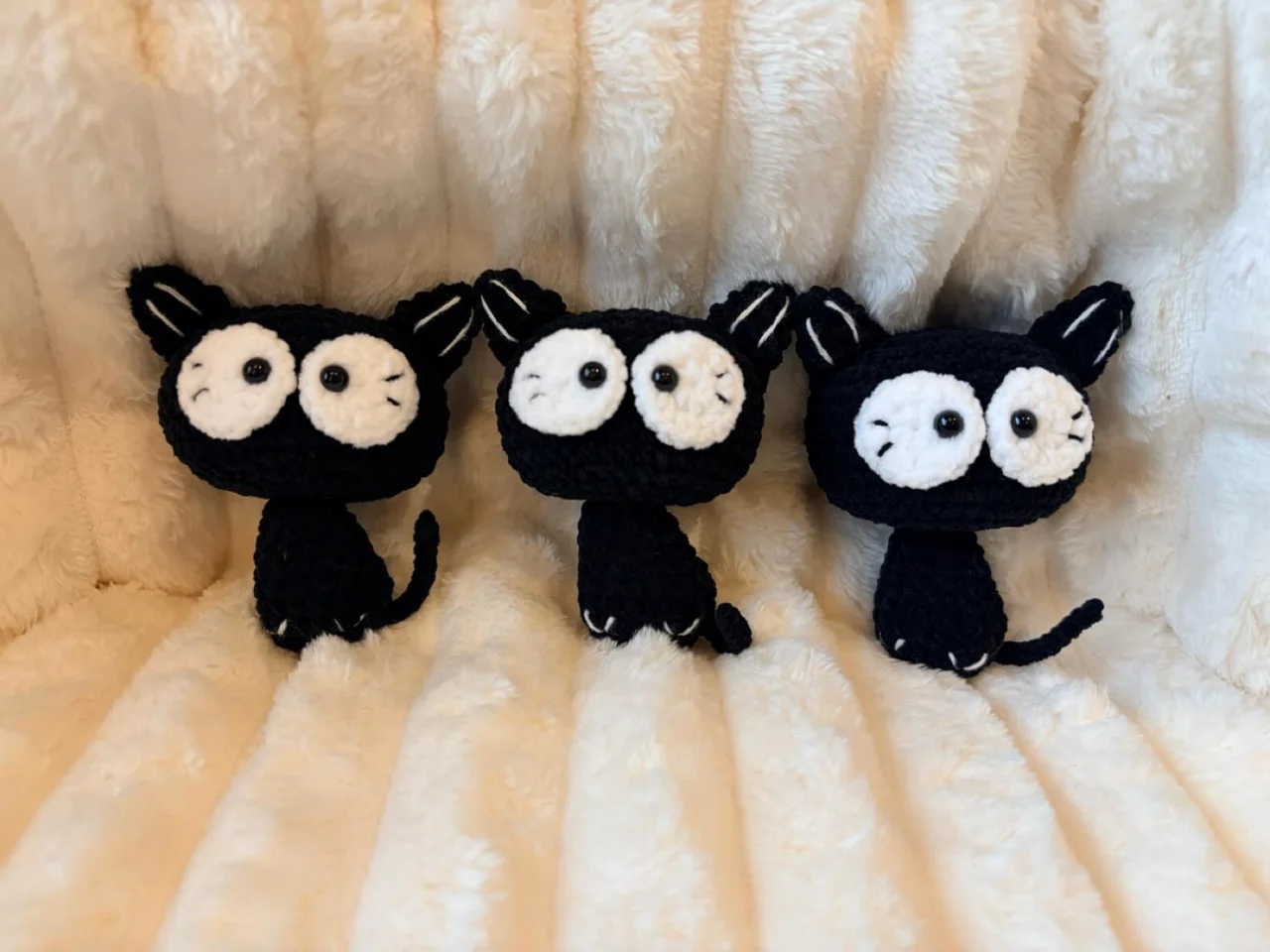 Three black crochet cats with large white eyes and stitched whiskers, sitting on a white blanket