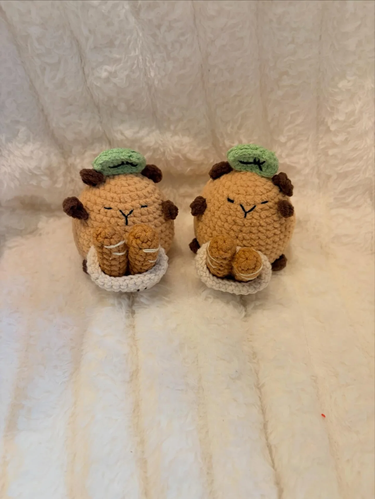 Two small crochet hedgehogs with brown spines, cream feet and green leaf hats, sitting on a white blanket